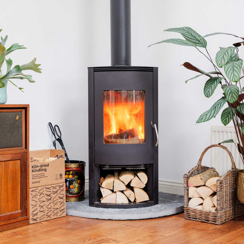 ECOFUEL Ash firewood-burning stove with fire burning inside, surrounded by ECOFUEL kindling box, basket of wood, and plants.