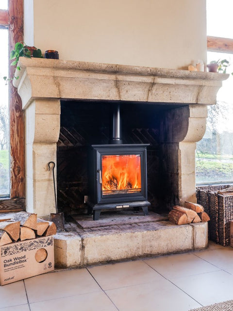 A black wood-burning stove with a bright orange fire, set inside a large stone fireplace. A cardboard box of 'Oak Wood BundleBox' firewood is visible on the left, and the room features beige tiled floors and a light-colored wall.