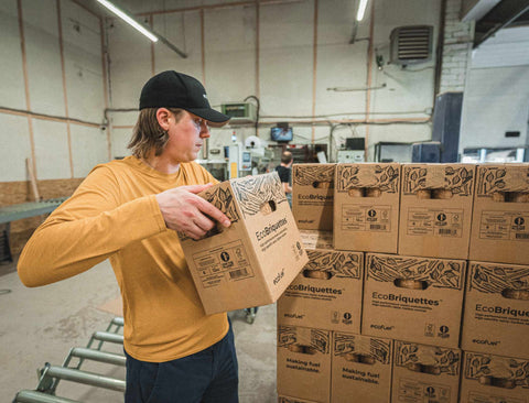 Janis in a warehouse holding an 'EcoBriquettes' box with stacked boxes in the background.