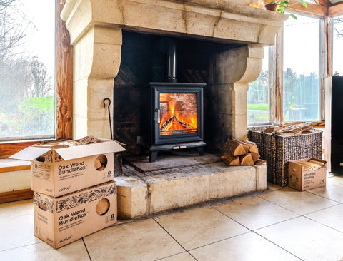 A black wood-burning stove with a visible fire sits inside a large stone fireplace. Cardboard boxes labelled ECOFUEL Oak Wood BundleBox are stacked on the floor to the left, and stacked firewood and baskets sit to the right in a rustic room with large windows.